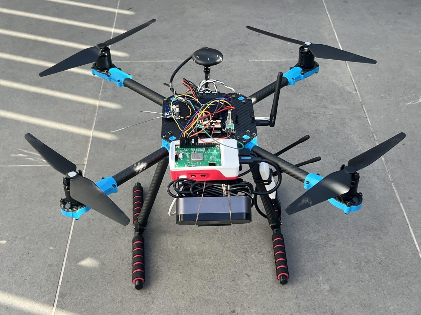 Guardian Eye montado por completo, visto de frente en tres cuartos: el mástil del GPS arriba, la placa de la Pixhawk 6X con su cableado, la Raspberry Pi 5 en su carcasa y la batería sujeta debajo, apoyado sobre el tren de aterrizaje rojo y negro