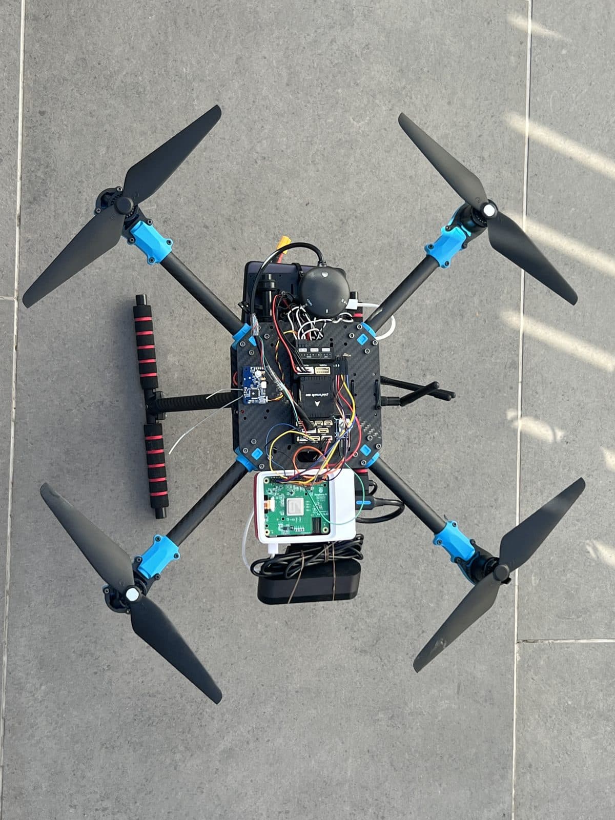 Vista cenital del dron completo: los cuatro brazos con motores y hélices, la placa central con la Pixhawk 6X y el domo del GPS, y la Raspberry Pi 5 con la batería en la bandeja inferior