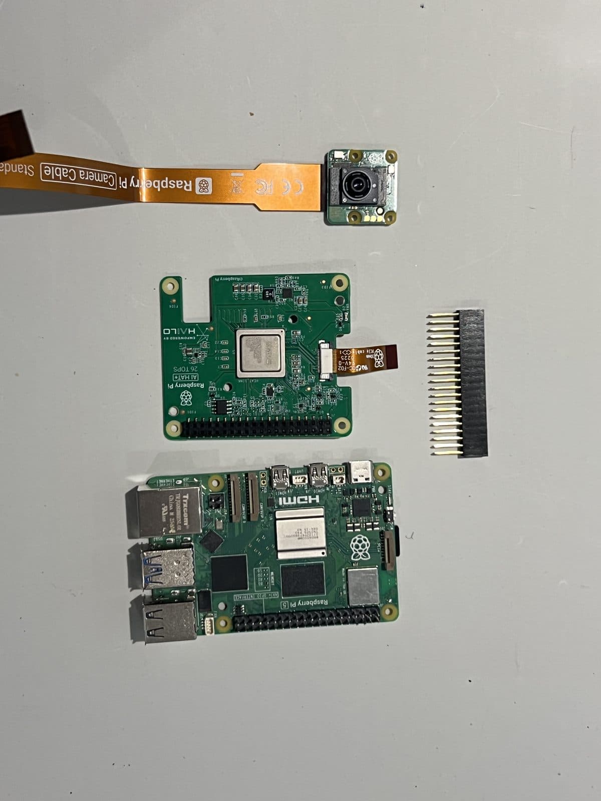 Cámara Raspberry Pi con su cable, el HAT Hailo-8 con su regleta extensora de GPIO y la Raspberry Pi 5, las tres piezas separadas antes de montar